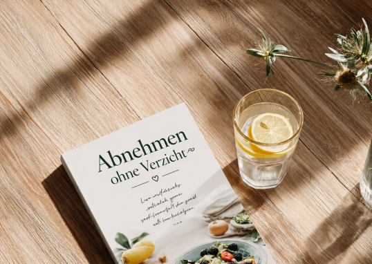 Inspirierendes Lifestyle-Bild zum Thema Abnehmen ohne Verzicht: Ein Rezeptbuch mit gesunden Mahlzeiten und ein Glas Zitronenwasser auf einem sonnigen Holztisch. Das Motiv symbolisiert eine ausgewogene Ernährung, gesunde Gewohnheiten und mehr Wohlbefinden im Alltag.
